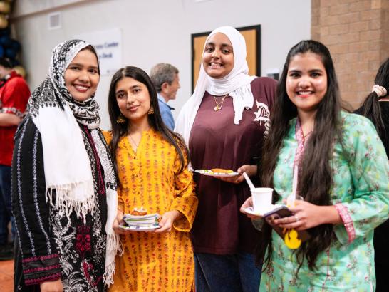 Four smiling international students posing together at an event, wearing traditional garments and hijabs while holding plates of food.