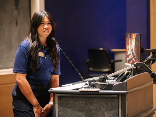 A woman wearing a navy blue polo stands in front of a microphone. 