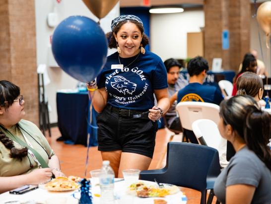 Student intern socializing at event with colorful blue and gold balloons in the foreground and backdrop