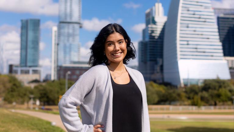 A student stands confidently with her hand on her hip in front of the Austin skyline