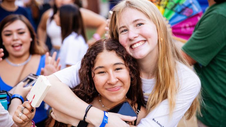 A close-up shot of two smiling young women hugging at a crowded outdoor block party. The woman on the right has blonde hair and is wrapping her arm around the shoulders of her friend, who has curly brown hair and is holding a smartphone.