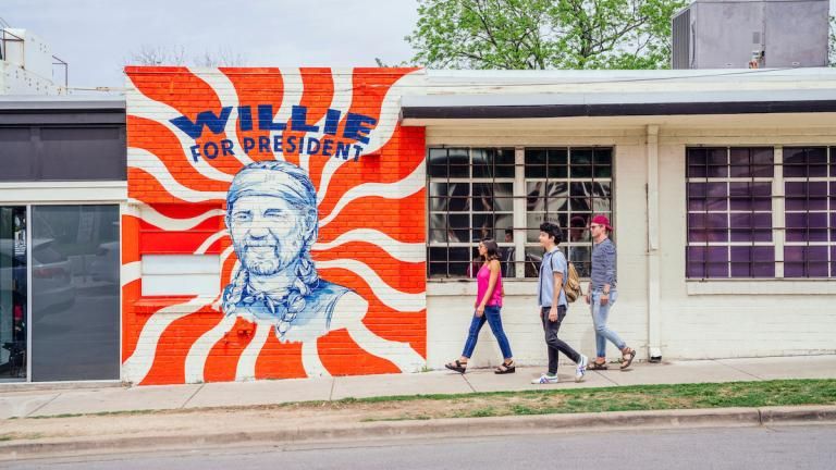 Three St. Ed's students walk down South Congress Avenue in front of a red and white mural showing an illustration of Willie Nelson that says "Willie for President"