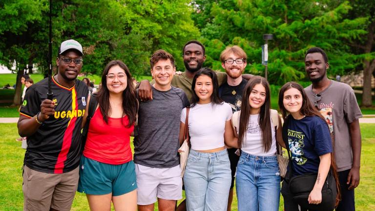 A diverse group of nine smiling students posing closely together on a green campus lawn with trees in the background.