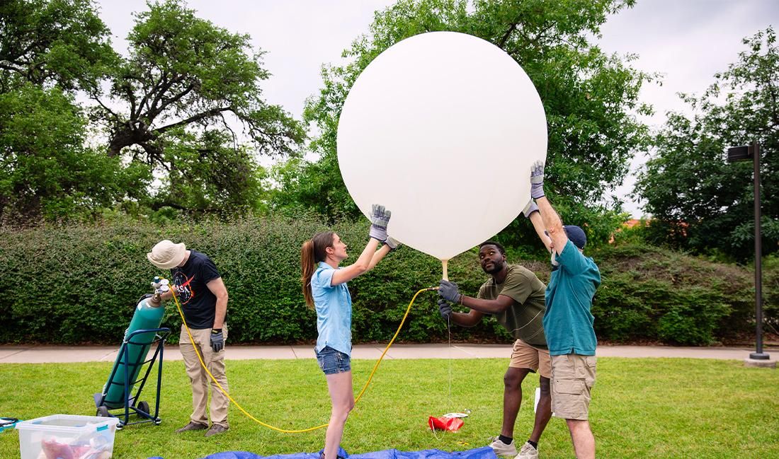 The image shows a group of four people outdoors, working together to inflate a large weather balloon. They are standing on a grassy area surrounded by trees and bushes. Two people are holding the balloon steady, while another person appears to be controlling the flow of gas from a tank connected to the balloon. All of them are wearing gloves, and one person is wearing a hat for sun protection. The scene seems to be part of a scientific or educational activity, likely related to an eclipse observation.