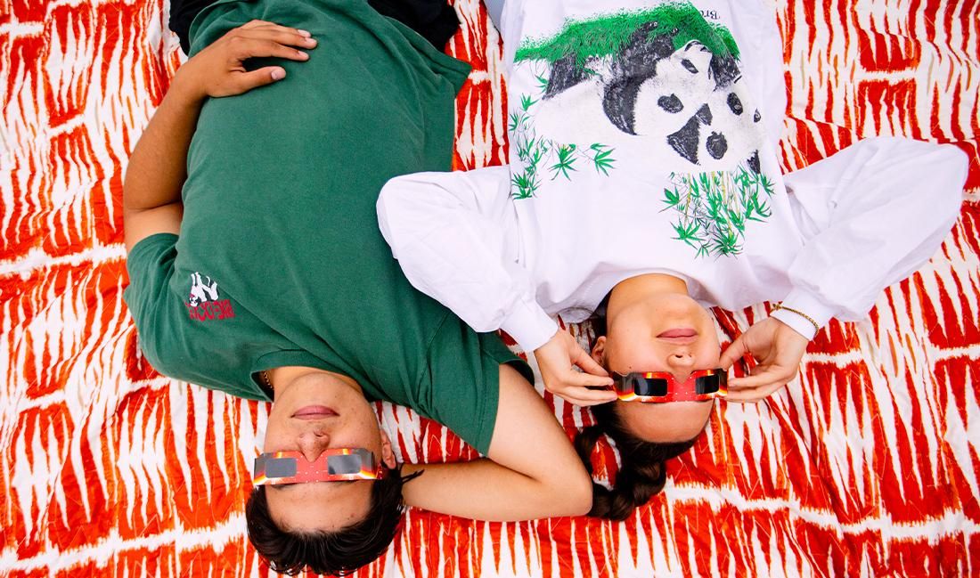 The image shows two people lying side by side on a brightly patterned red and white blanket, looking up at the sky through eclipse glasses. One person is wearing a green T-shirt, while the other is dressed in a white long-sleeve shirt featuring pandas. Both individuals appear relaxed and ready to watch a solar eclipse, with their hands adjusting their glasses. The scene captures a casual and enjoyable moment of shared experience during an eclipse viewing.