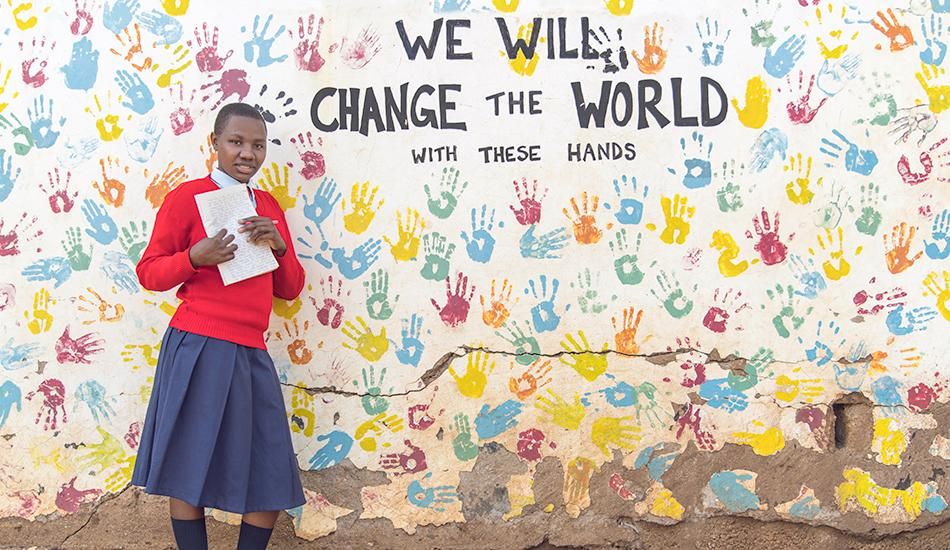 Student standing in front of a wall painting that says we will change the world. 