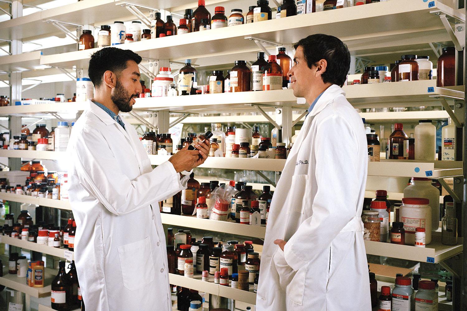 Diego Saldana and Professor Santiago Toledo Carrion wear white lab coats and stand by shelves of bottles of chemicals and compounds in a lab.