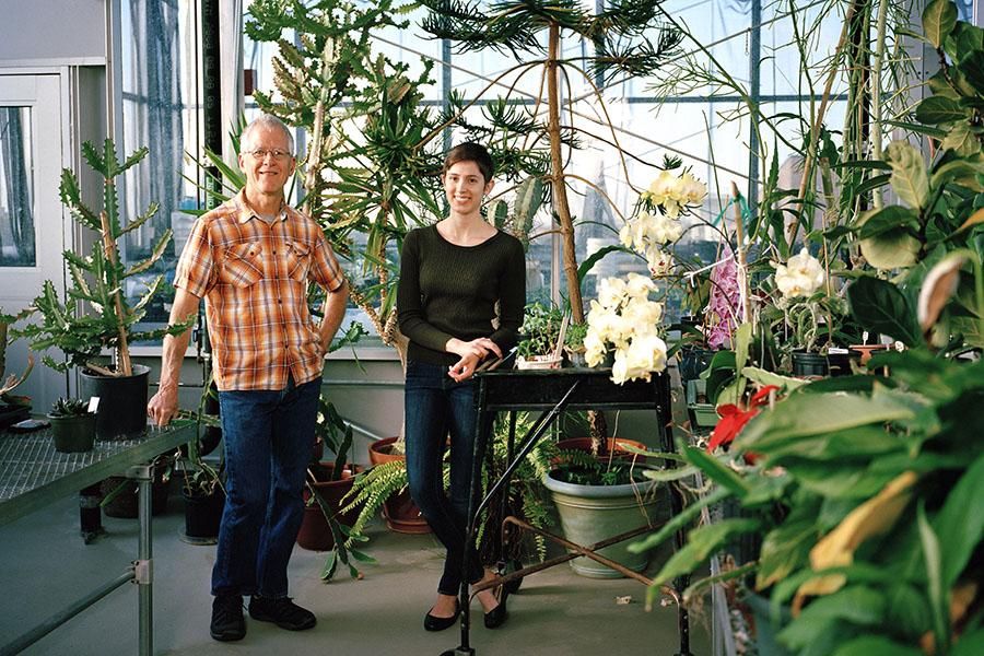 Professor Charles Hauser and Jacquelyn Turcinovic stand in the greenhouse, surrounded by greenery and flowers.