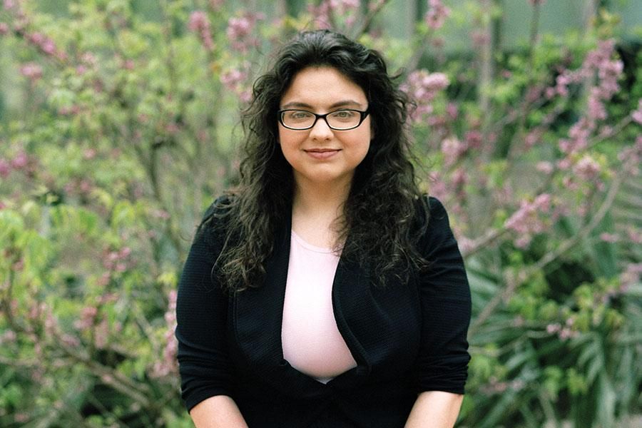 Ciara Crochet wears a light pink shirt, a black blazer and glasses, and sits in front of greenery and flowers.