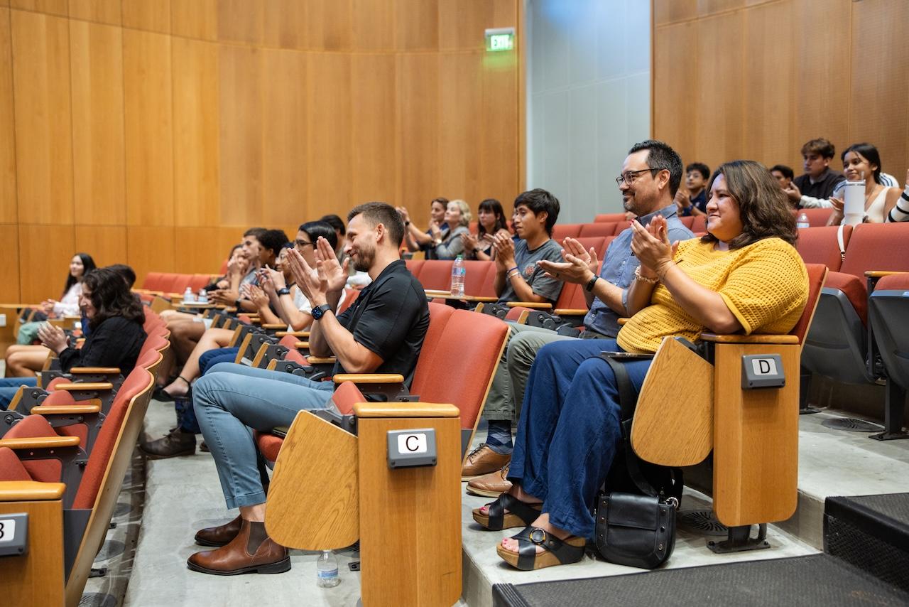 A group of college students sit in an auditorium and clap. 