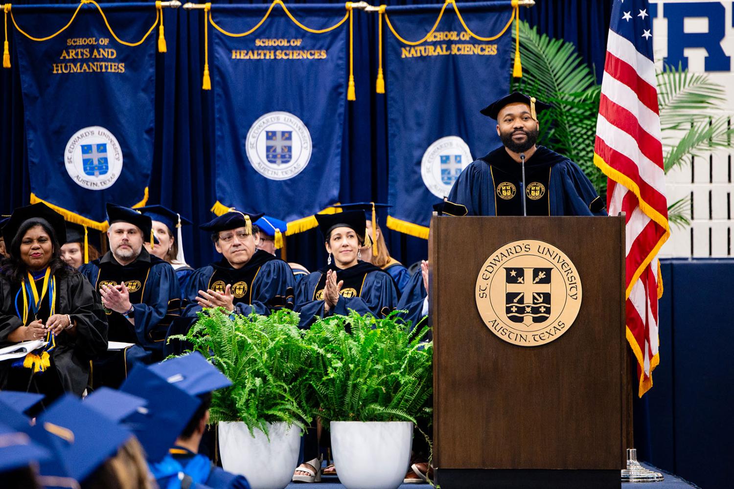 Members of the board and cabinet at St. Edward's University, dressed in commencement regalia, sit and clap behind Linus Akanoh, Chair of the Board of Trustees, who speaks in front of a podium on stage at the university's commencement ceremony.